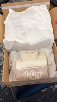 Brevet krem- og beige strikkegenser med teksten "LeGer", brettet sammen i en pappeske foret med hvitt krøllet silkepapir på en mønstret overflate.