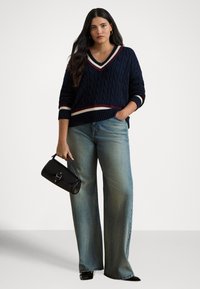 Ženska nosi mornarsko modri kabelsko pleten pulover z V-izrezom z belimi in bordo črtami, široke kavbojke, črne pete in drži črno torbico.