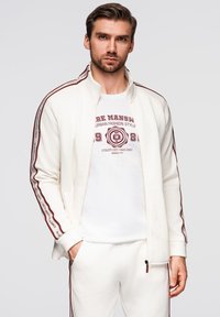 Chaqueta blanca con cremallera y rayas laterales en color burdeos, de textura suave. Llevada sobre una camiseta blanca con texto gráfico en rojo. Pantalones blancos a juego.
