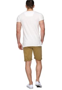 Biały t-shirt z krótkim rękawem o luźnym kroju, zestawiony z khaki szortami z podwiniętymi nogawkami. Model w białych trampkach, widziany od tyłu.