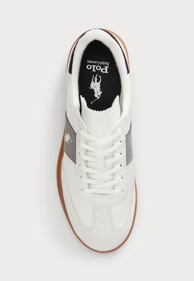 Sapatilha branca com risca cinza, parte superior em couro e têxtil, atacadores brancos planos e sola de borracha castanha. Apresenta logo na língua e na lateral.