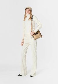 Mono color crema con cuello alto, piernas acampanadas y costuras contrastantes, combinado con un bolso bandolera beige y una gorra de color claro.