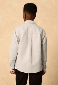Light gray button-up shirt with long sleeves, single back pleat, and buttoned cuffs, worn with dark pants. Simple, clean design.
