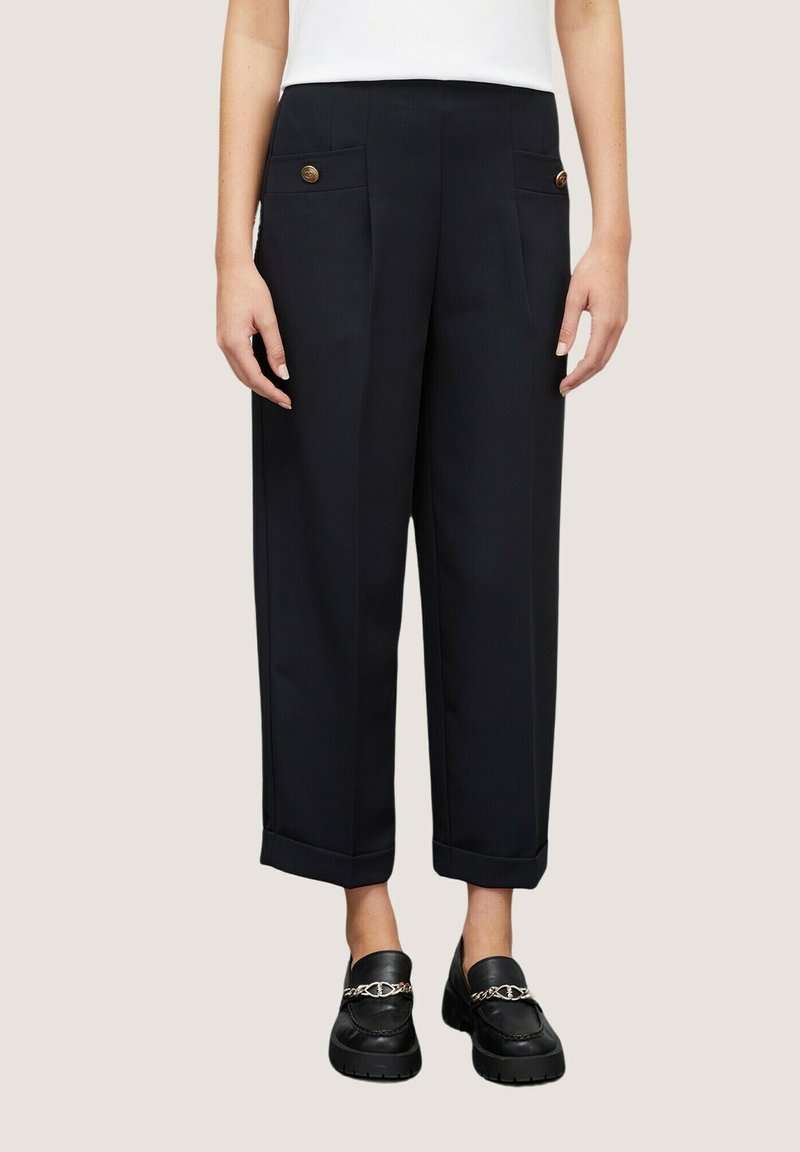 Zwarte wijdgesneden broeken met voorplooien, zijzakken en gouden knopen. Omgeslagen zoom en tailor-made ontwerp. Draagt met zwarte loafers.