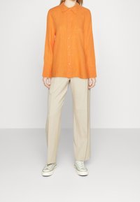 Camicia arancione in lino con colletto, abbinata a pantaloni beige a gamba larga e sneakers verde chiaro. Tessuto liscio, vestibilità rilassata.