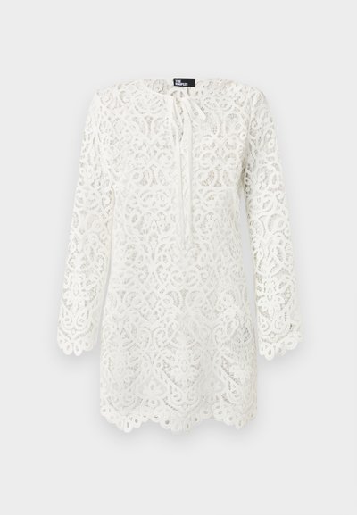 Blouse blanche en dentelle à manches longues avec motifs floraux complexes, bords festonnés et petit nœud au col.