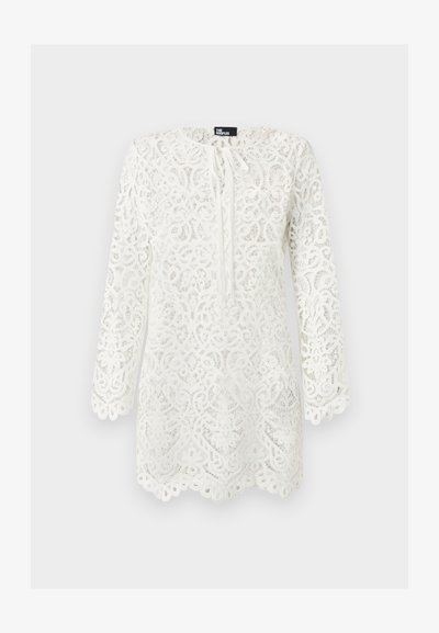 Blouse blanche en dentelle à manches longues avec motifs floraux complexes, bords festonnés et petit nœud au col.