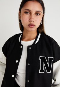 Veste de style varsity noire et blanche avec fermeture à boutons, ornée d'un grand patch blanc en forme de 'N' sur la poitrine et d'un col côtelé, portée sur un top blanc.