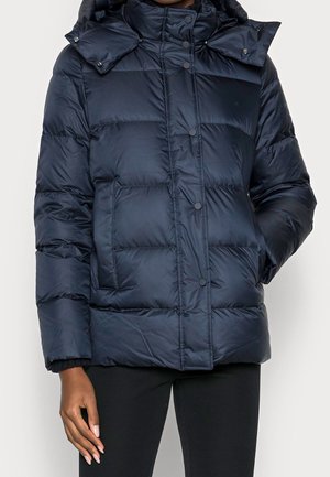 Vrouw draagt een marineblauwe gewatteerde jas met drukknopen en grote voorzakken, gecombineerd met een zwarte broek.