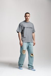 Noor mees seisab seljas hall suurendatud "AY STUDIOS" t-särgi, rebenenud heledates sinistes teksades ja valgete ning siniste spordijalanõudega valgel taustal.