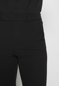 Musta, kapealahkeiset housut, sileä pinta, vyölenkit ja etutaskut, joissa on tavallinen vyötärönauha ilman näkyviä koristeita.