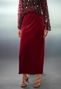 Falda maxi de terciopelo burdeos combinada con una blusa transparente adornada con lentejuelas, que presenta mangas largas y un corte holgado, complementada con tacones con tiras.