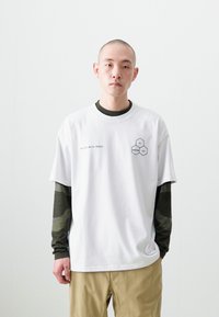 White cotton t-shirt with short sleeves, featuring a hexagon graphic and text "FRIENDS WITH TREES." Layered over a long-sleeve green camo shirt.
