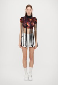 Top corto negro con un patrón abstracto colorido; cuenta con flecos largos negros. Combinado con una mini falda blanca y zapatillas blancas.