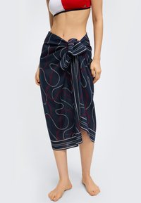 Γυναίκα που φοράει ναυτικό σαρόνγκ με κόκκινα και λευκά σχέδια αλυσίδας, δεμένο στη μέση, συνδυασμένο με κόκκινο και λευκό κοντό τοπ.