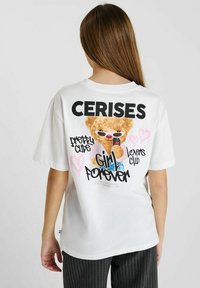 T-shirt en coton blanc avec des impressions graphiques colorées, y compris un ours en cartoon, le texte "CERISES" et des cœurs. Coupe décontractée, manches courtes.