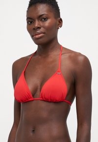 Haut de bikini triangulaire rouge en tissu lisse, avec de fines bretelles et des détails froncés, arborant un petit logo près de la couture.