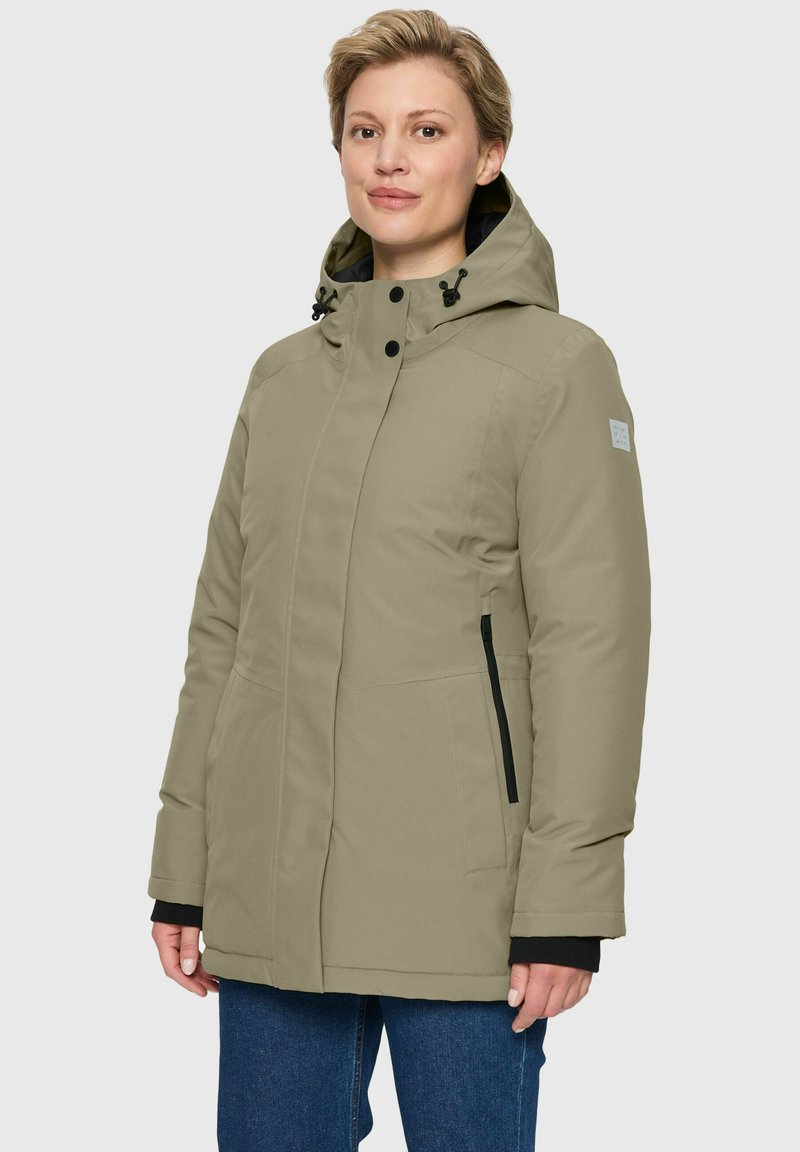 Eine Frau trägt eine beige Winterjacke mit Kapuze und schwarzen Reißverschlusstaschen und steht vor einem schlichten hellen Hintergrund.