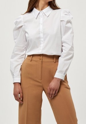 Chemise blanche à boutons avec manches bouffantes, associée à un pantalon beige taille haute. La chemise présente un col classique et un tissu lisse.