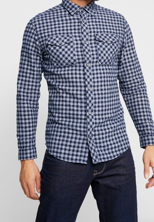 Mann trägt ein blau-weiß kariertes Flanellhemd mit geknöpften Brusttaschen und dunkelblaue Jeans vor einem einfarbigen Hintergrund.