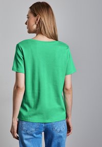 Zelené bavlněné tričko s krátkým rukávem a širokým výstřihem, spárované se světle modrými džíny. Tkanina vypadá měkce a má hladkou texturu.