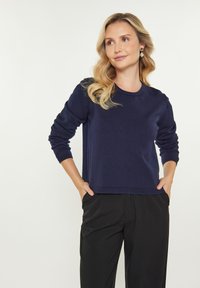 Pulover tricotat bleumarin cu guler rotund, mâneci lungi și croială lejeră, purtat cu pantaloni negri pe măsură, cu mâinile în buzunare.