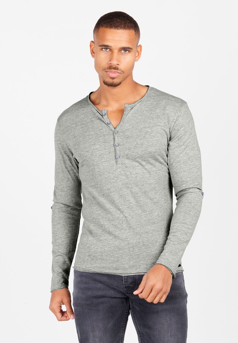 Langärmliges graues Henley-Shirt mit einem V-Ausschnitt, verfügt über vier Knöpfe und besteht aus einem weichen, strukturierten Stoff. Getragen mit dunklen Jeans.