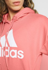 Sweat à capuche rose avec une poche kangourou à l'avant, une capuche à cordon de serrage et un logo Adidas blanc. Tissu doux avec une texture lisse et une coupe décontractée.