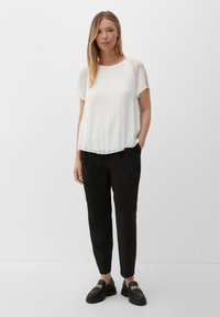 Blouse blanche plissée avec des manches courtes transparentes, associée à un pantalon noir tapered et des mocassins noirs épais avec des accents de chaîne dorée.