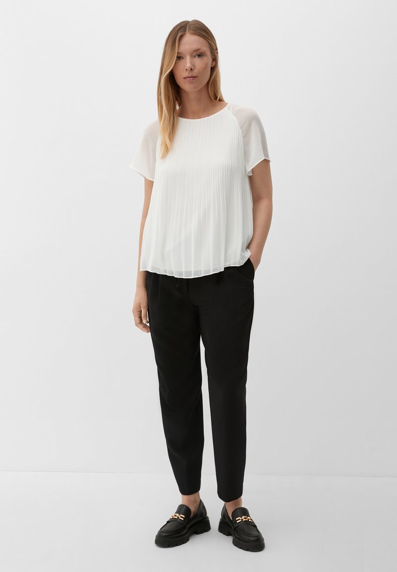 Blouse blanche plissée avec des manches courtes transparentes, associée à un pantalon noir tapered et des mocassins noirs épais avec des accents de chaîne dorée.