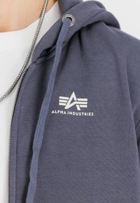 Γκρίζα φούτερ με φερμουάρ από μαλακό ύφασμα, με λευκό λογότυπο που έχει γεωμετρικά σχήματα και μεταλλική άκρη στην κλωστή.