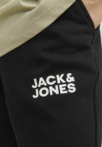 Črne trenirke z gladko teksturo imajo bel logo "JACK & JONES" na levem stegnu, v kombinaciji s svetlo obarvanim zgornjim delom.