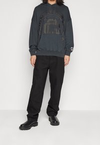 Dunkelblauer Kapuzenpullover mit Logo-Grafik, Fronttasche und verstellbarem Hoodie, kombiniert mit locker sitzenden schwarzen Hosen und schwarzen klobigen Stiefeln.