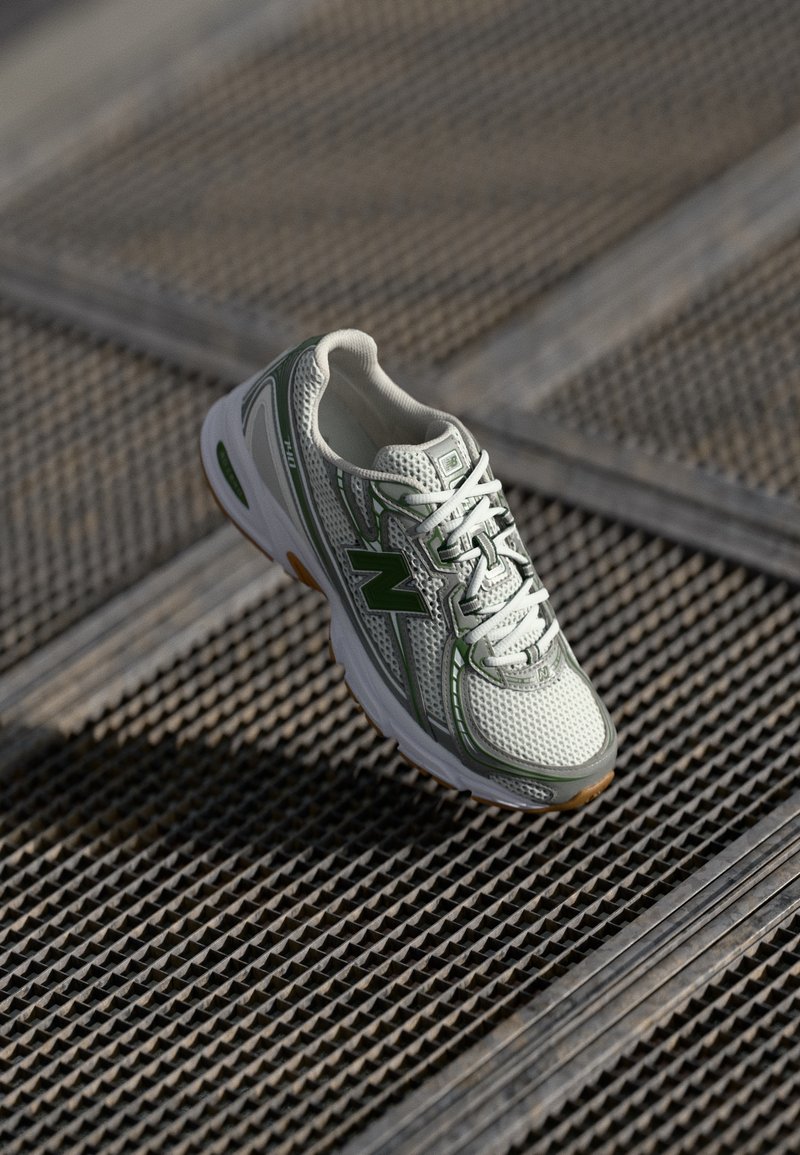 Chaussure de running New Balance blanche et verte avec une tige en mesh, des lacets blancs et une semelle en caoutchouc sur une surface métallique texturée.
