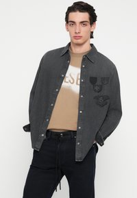 Σκούρο γκρι πουκάμισο από τζιν με κουμπιά μπροστά, με τρεις μαύρες επιδιορθώσεις στο στήθος. Φορεμένο πάνω από μία μπεζ γραφική μπλούζα. Σκούρο παντελόνι.