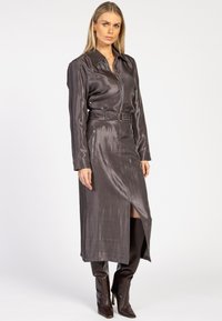 Vrouw in een glanzende donkergrijze midi-jurk met ceintuur, met een voorknijp en lange mouwen, gecombineerd met donkerbruine laarzen tot aan de knie.