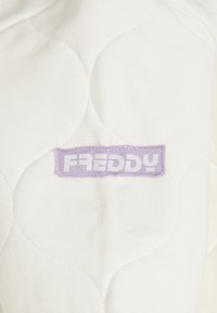 Tela blanca acolchada con un patrón de costuras, con una etiqueta rectangular morada que muestra la palabra "FREDDY" en letras capitales blancas.