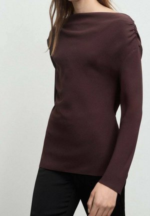 Femme portant un haut ajusté à manches longues de couleur bordeaux foncé avec des détails froncés aux épaules et un pantalon noir.