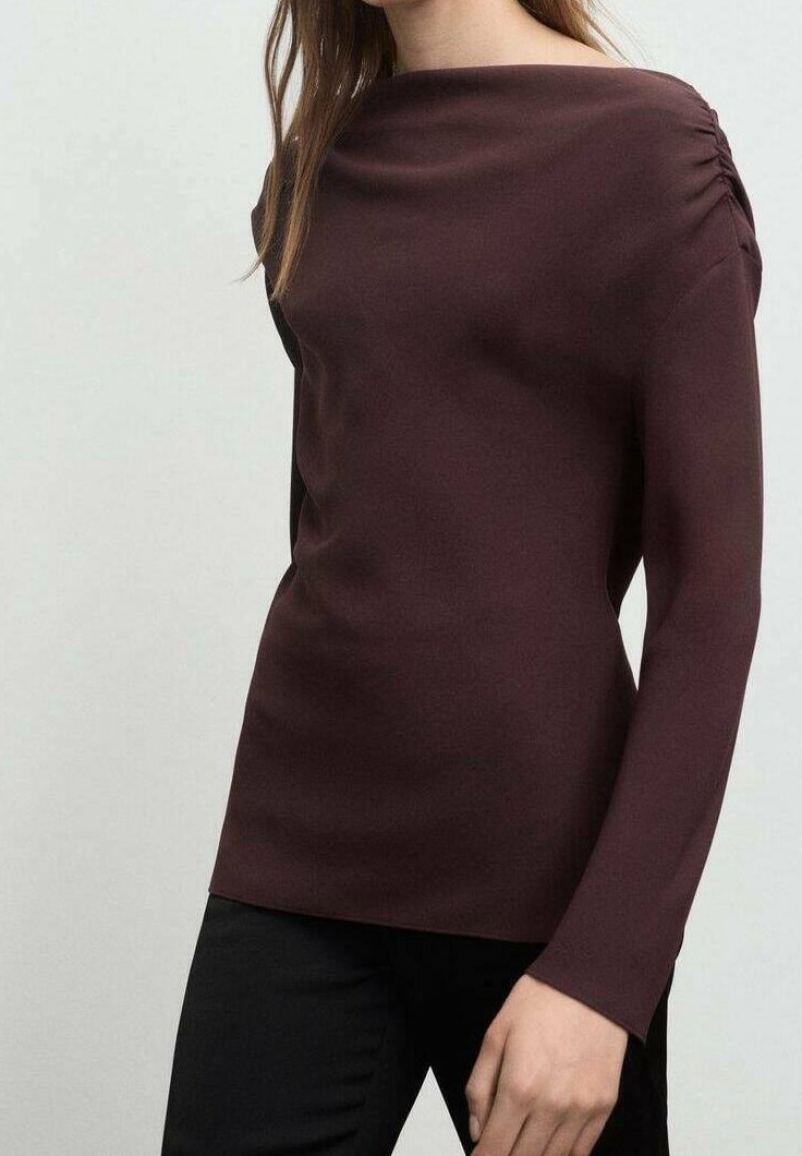 Femme portant un haut ajusté à manches longues de couleur bordeaux foncé avec des détails froncés aux épaules et un pantalon noir.