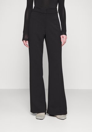 Frau trägt hoch taillierte, schwarze Hose mit weitem Bein, kombiniert mit einem enganliegenden schwarzen Langarmoberteil und hellen Schuhen, steht vor einem schlichten Hintergrund.