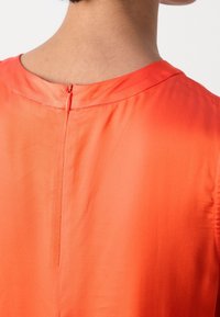Blusa arancione con una morbida texture in satin, caratterizzata da un collo rotondo e una cerniera discreta sul retro per la chiusura.