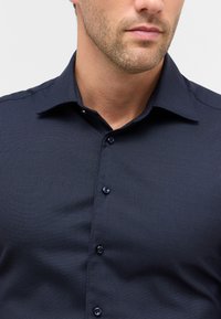 Uomo che indossa una camicia da uomo button-down blu scuro con colletto strutturato e leggera barba incolta sul viso, con uno sfondo semplice.