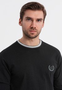 Schwarzes langärmliges Hemd mit einem grau gestreiften Kragen, ausgestattet mit einem kleinen gestickten Logo auf der Brust. Glattes Baumwollgewebe.