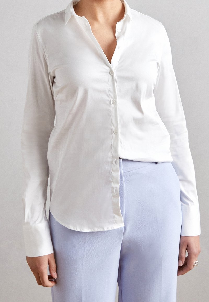 Camicia bianca con bottoni, maniche lunghe, tessuto liscio, colletto classico e orlo curvo, abbinata a pantaloni azzurri chiari.