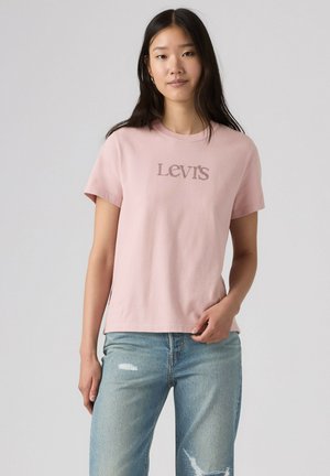 Young woman wearing a light pink Levi's t-shirt and light blue ripped jeans standing against a plain gray background.