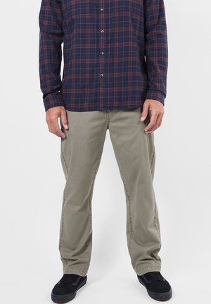 Homme portant un pantalon beige, des baskets noires et une chemise à carreaux bleu foncé et rouge boutonnée, debout devant un fond uni.
