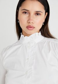 White button-up blouse with a high ruffled collar, featuring front button closure and subtle gathers at the shoulders. Smooth fabric texture.
