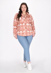 Koszula z długim rękawem, zapinana na guziki w kolorze koralowym z białymi wzorami geometrycznymi, zestawiona z jasnoniebieskimi obcisłymi dżinsami i białymi trampkami. Gładka faktura materiału.