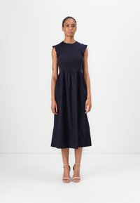 Marineblaues Kleid mit tailliertem Oberteil, ausgestelltem Rock und Rüschendetails an den Schultern. Glatte Textur, Midi-Länge, kombiniert mit nude farbenen High Heels.