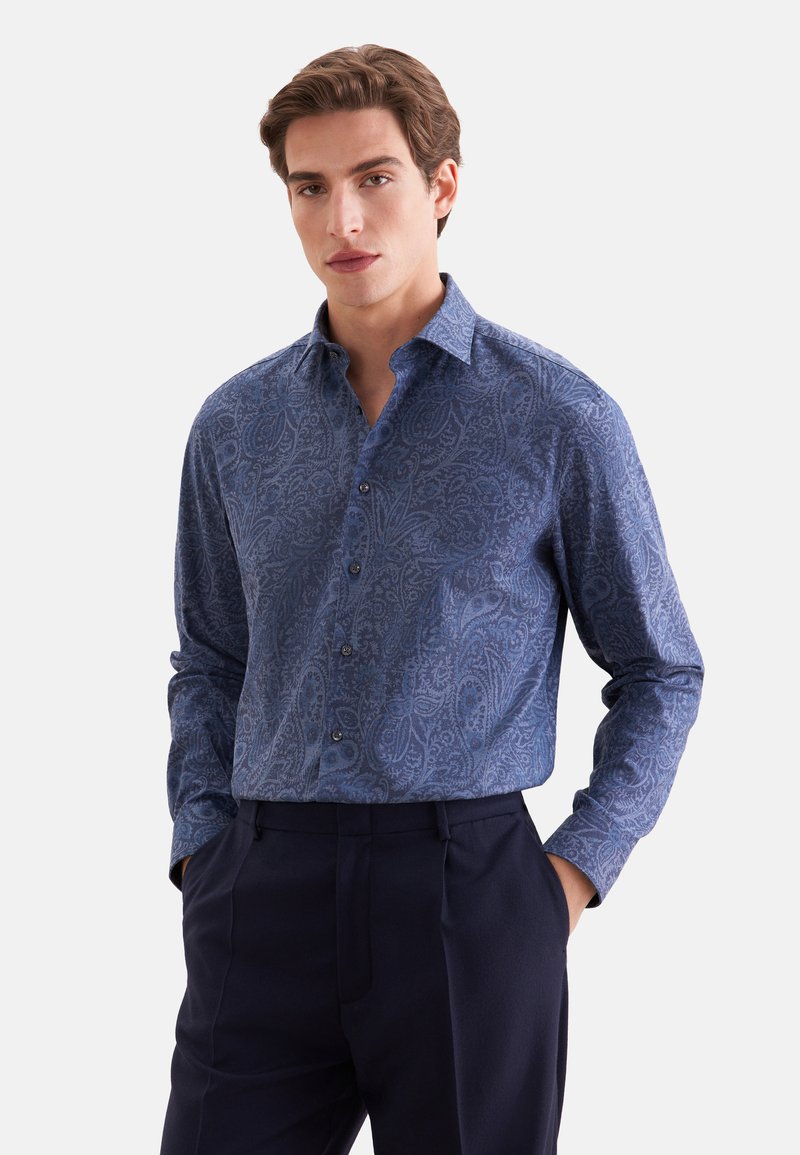 Homme aux cheveux châtain clair portant une chemise bleue à motifs paisley rentrée dans un pantalon bleu marine foncé, mains dans les poches.